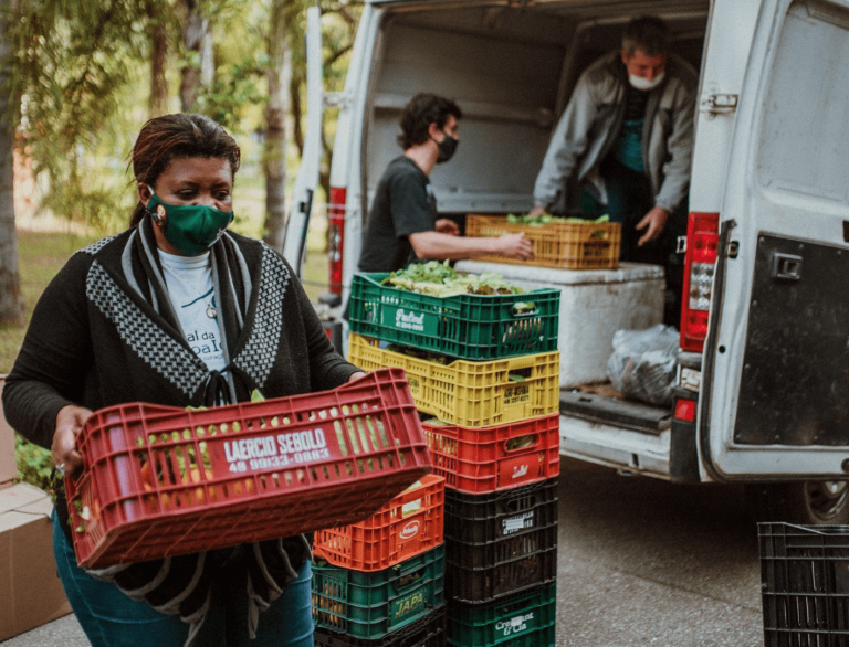 Imagem mostra uma mulher com máscara carregando uma caixa de alimentos frescos enquanto outras pessoas descarregam mais caixas de um veículo. A cena representa ações de apoio comunitário e distribuição de alimentos, simbolizando a atuação da filantropia empresarial no enfrentamento de desigualdades sociais, promovendo segurança alimentar, bem-estar e desenvolvimento local.