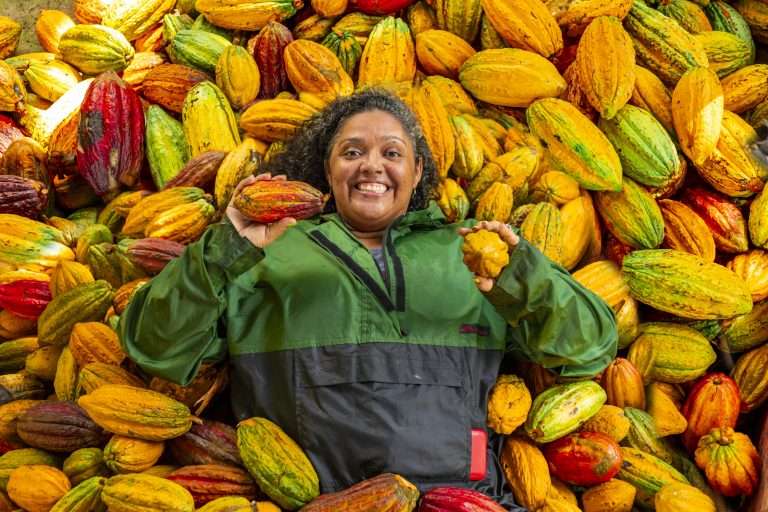 Mulher sorridente segurando frutos de cacau em meio a uma grande quantidade de vagens coloridas, representando o protagonismo de produtores locais em cadeias sustentáveis. A imagem ilustra como o investimento de impacto no Brasil pode promover o desenvolvimento econômico aliado à conservação ambiental, alinhando-se a práticas ESG ao valorizar cadeias da sociobiodiversidade e impulsionar negócios que geram impacto social positivo em territórios diversos.
