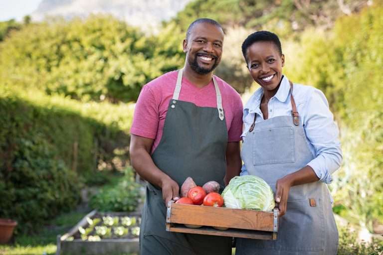 Casal sorridente com aventais segurando uma caixa de legumes frescos em uma horta, simbolizando produção sustentável e economia local. A imagem representa como empresas, especialmente pequenos negócios, podem contribuir ativamente com os Objetivos de Desenvolvimento Sustentável (ODS) da Agenda 2030, promovendo inclusão social, segurança alimentar, trabalho digno e práticas ambientalmente responsáveis.