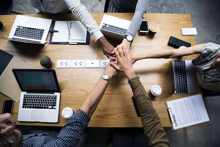 Vista superior de uma mesa de reunião com laptops, documentos e cinco pessoas unindo as mãos no centro, simbolizando união, compromisso e trabalho em equipe. A imagem representa a responsabilidade social corporativa (RSC) como um valor que conecta profissionais em torno de propósitos comuns — promovendo ações empresariais alinhadas ao bem-estar coletivo, à ética nos negócios e ao desenvolvimento sustentável.