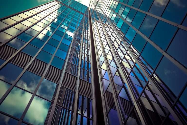Imagem de um arranha-céu moderno com fachada de vidro refletindo o céu e as nuvens, vista de baixo para cima, simbolizando crescimento, visão de futuro e atuação corporativa. A cena representa o papel das empresas na promoção da filantropia estratégica, em que ações sociais são alinhadas ao propósito institucional e aos objetivos de negócio, ampliando o impacto positivo e o valor gerado para a sociedade.