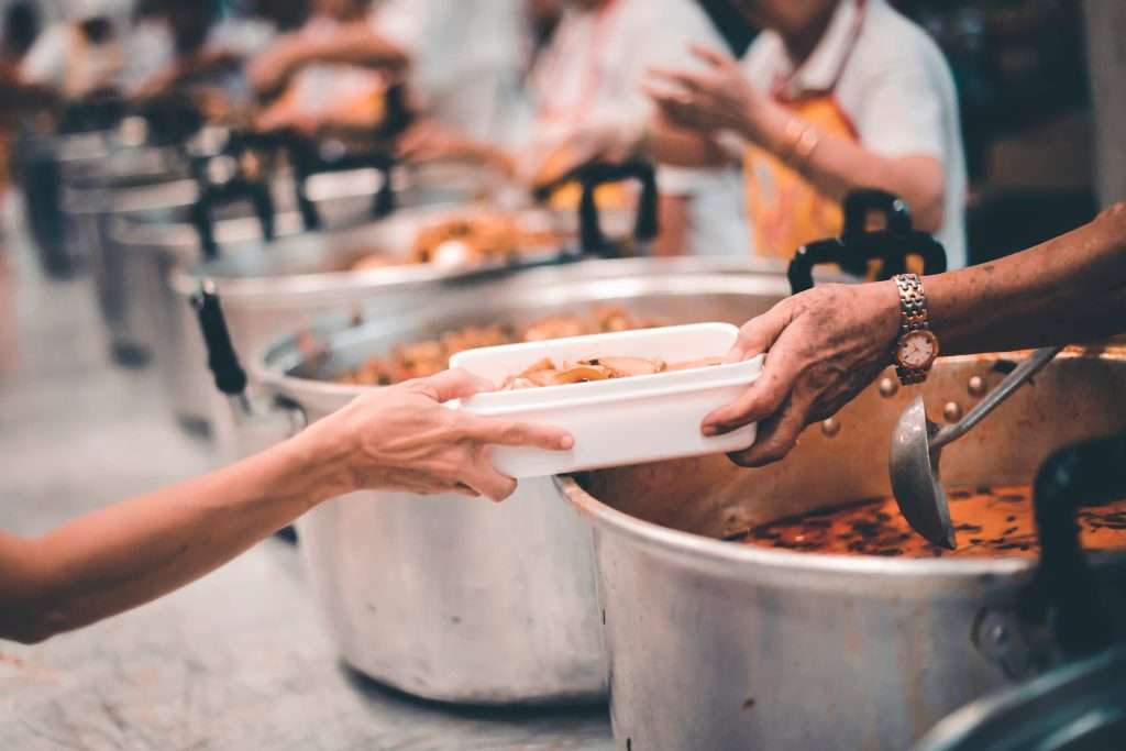Pessoa entrega uma marmita com comida a outra em um ambiente de cozinha comunitária, com grandes panelas ao fundo e outras pessoas servindo alimentos. A imagem retrata uma ação solidária típica de iniciativas promovidas por Organizações da Sociedade Civil (OSCs), que atuam no combate à fome, na redução das desigualdades e na promoção da cidadania.