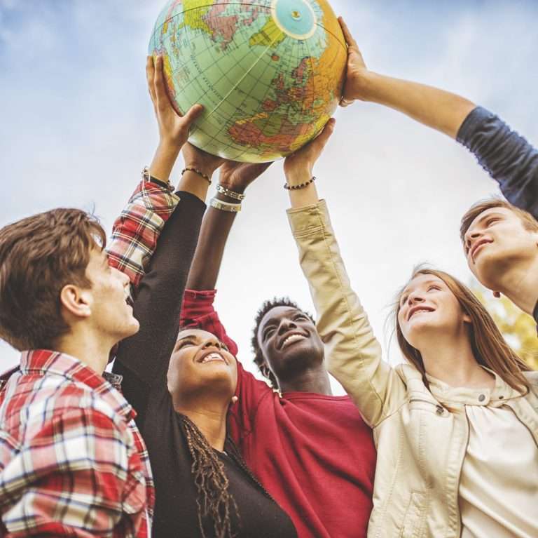 Grupo diverso de jovens segurando um globo terrestre juntos, com expressão de entusiasmo e cooperação, ao ar livre em um dia claro.
